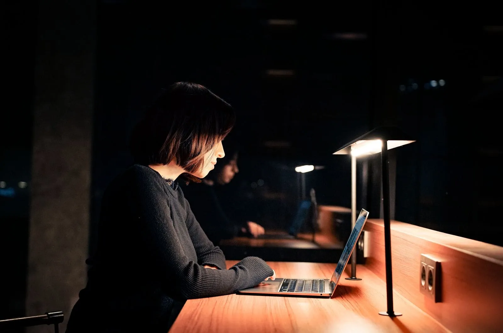 A woman at a desk in the evening, lamplight on, laptop open