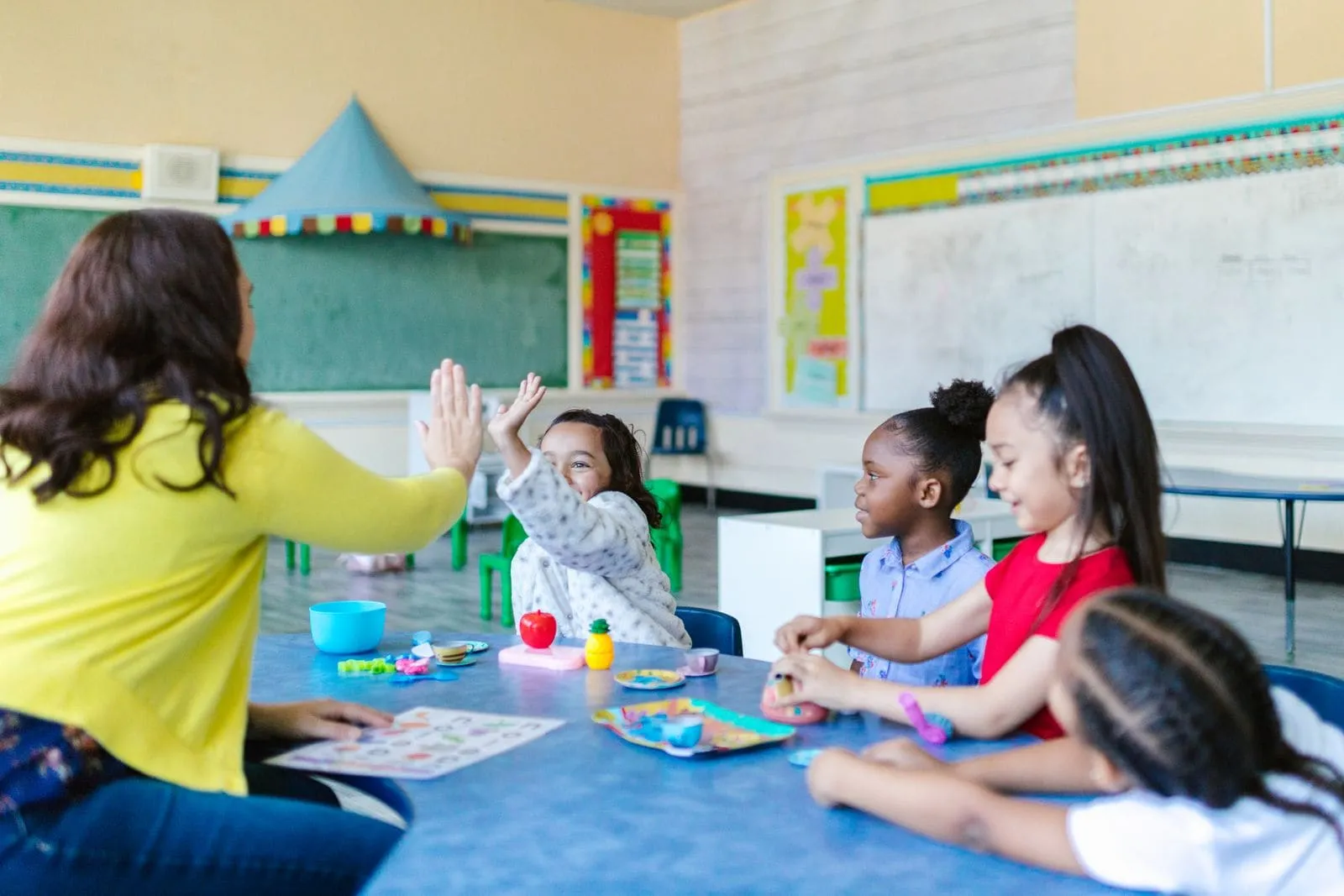 A teacher high-fives a young student at a classroom table while three other children look on